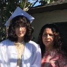 Woman in white graduation gown and cap, wearing glasses standing next to a woman with curly hair.