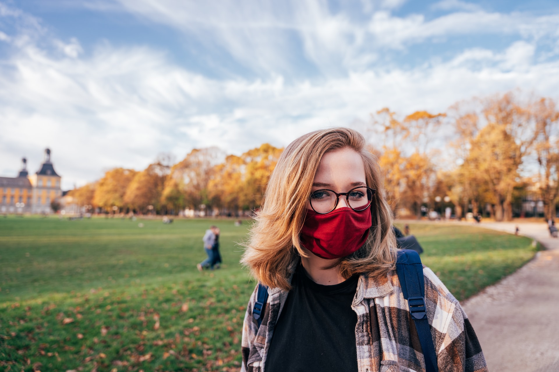 Woman with short blonde hair, standing in a park. Wearing a red cloth mask and a checkered flannel.