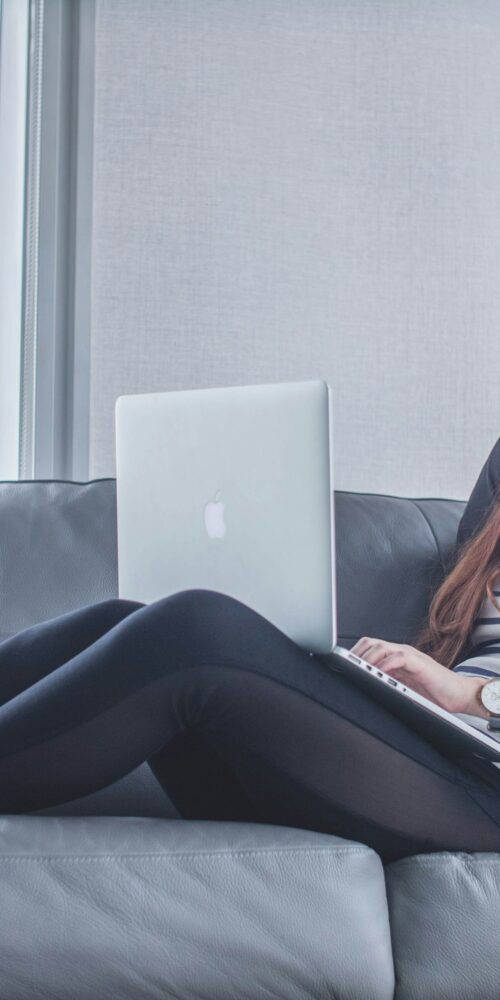 Woman in striped shirt using a laptop on a gray sofa, leaning against black pillows.