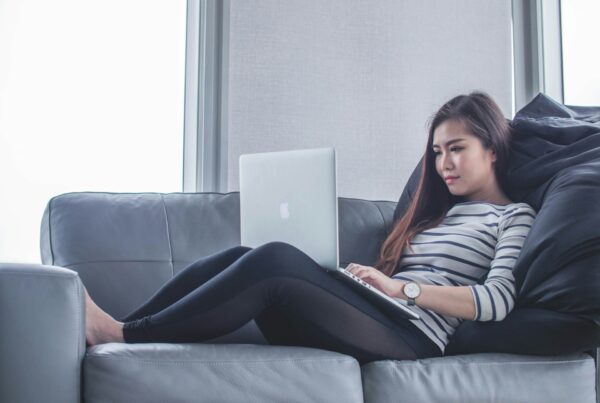 Woman in striped shirt using a laptop on a gray sofa, leaning against black pillows.